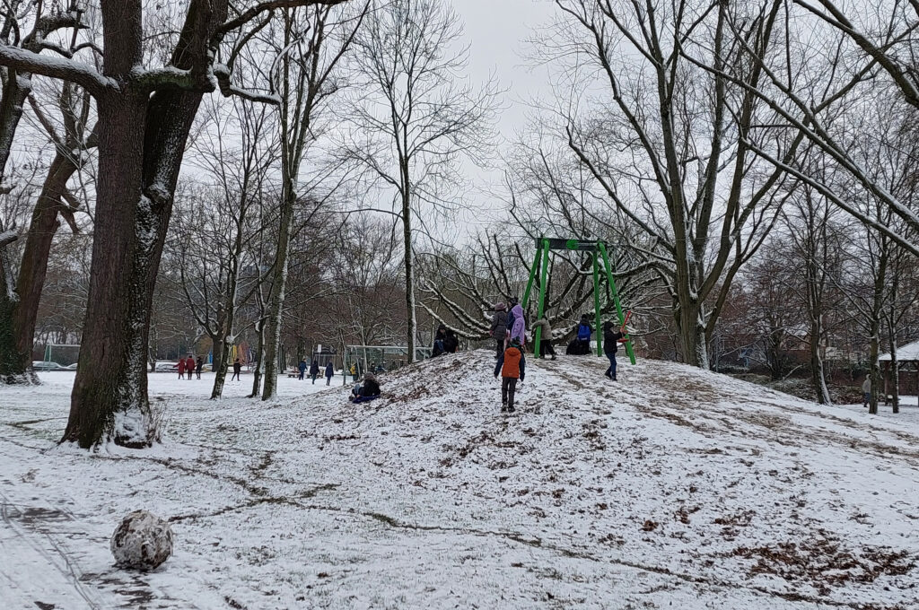 Zu sehen sind ein kleiner verschneiter Hügel, kahle Bäume bei grauem Wetter und ein paar spielende Kinder