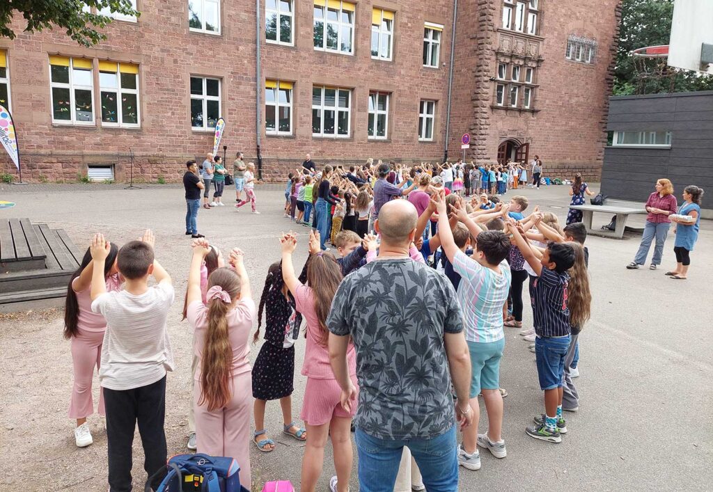 Vor dem Schulgebäude steht eine lange Schlange von Kindern, die sich zu zweit gegenüberstehen und mit erhobenen Armen ein Spalier bilden. Am Rand stehen Lehrkräfte, Erzieher*innen und Eltern und alle warten auf die Viertklässler*innen.