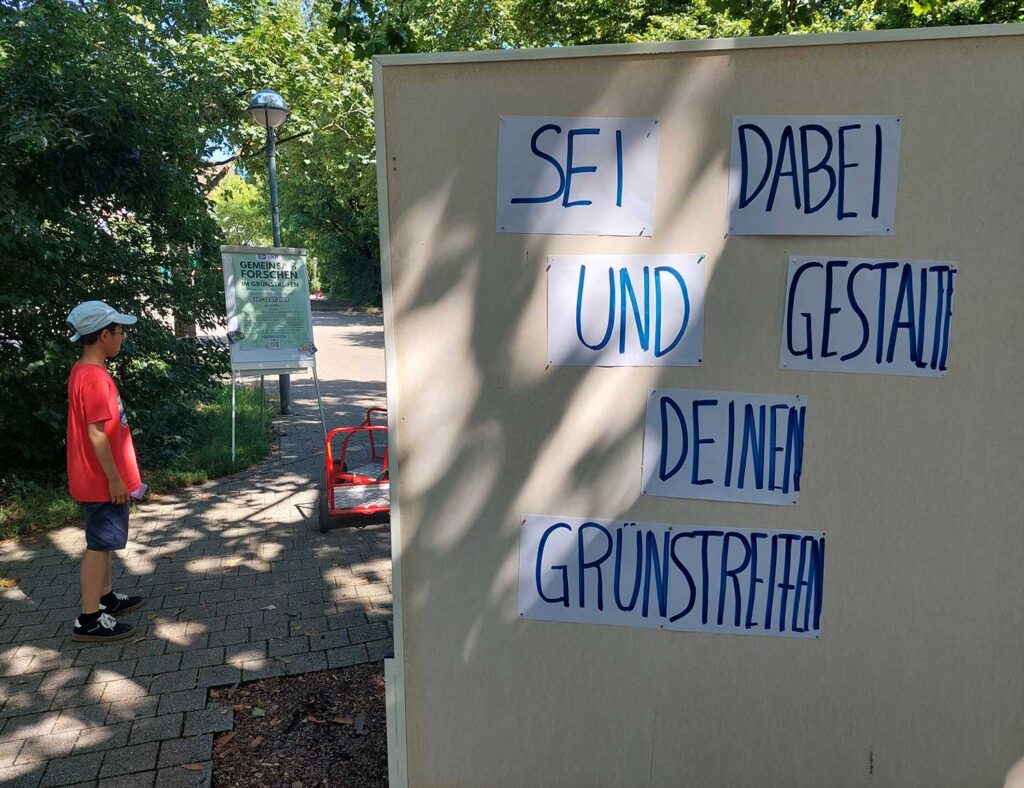 Ein Junge steht draußen im Grünen links im Bild und schaut auf ein Kinderfahrzeug, rechts sieht man eine Pinnwand mit der Aufforderung „Sei dabei und gestalte deinen Grünstreifen“.