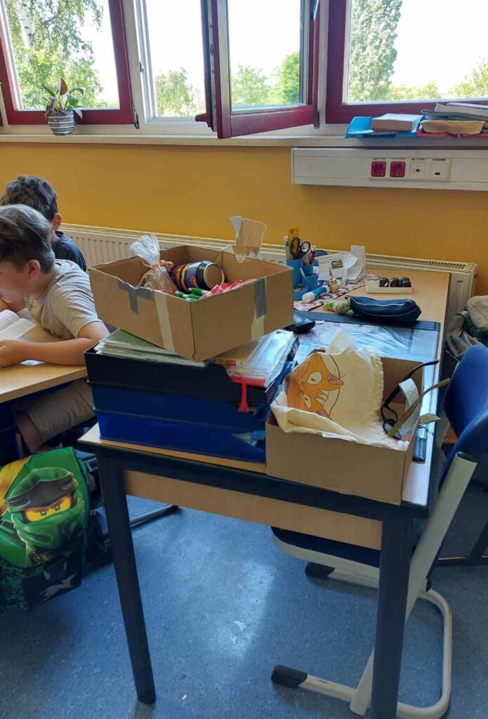 Wir sehen ein Klassenzimmer mit offenen Fenstern und am linken Rand zwei Schüler. Im Vordergrund steht der Tisch der Lehrerin mit Unterlagen und einem offenen Päckchen.