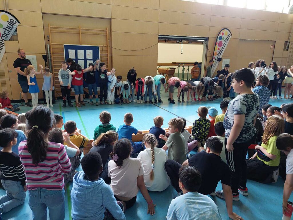 Im Vordergrund sieht man Kinder aus dem Publikum, die auf dem Boden der Sporthalle sitzen, im Hintergrund verbeugt sich eine Reihe von über 20 Kindern, die etwas vorgeführt bzw. moderiert haben.