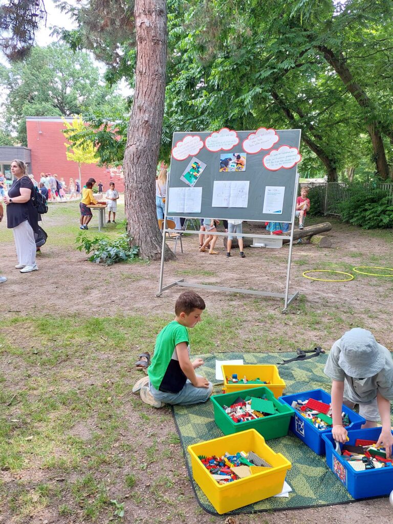 Im Vordergrund sitzen zwei Kinder im Schatten auf einer Picknick-Decke und bauen mit Lego, in der Mitte des Fotos steht eine Tafel mit Zitaten, Fotos und Gemälden aus dem Projekt, im Hintergrund sieht man mehr Eltern und Kinder auf dem Schulhof.