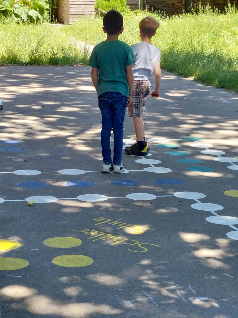 Zwei Jungs stehen mit dem Rücken zur Fotografin auf weißen Spielfeldern des auf dem Schulhof aufgemalten „Mensch ärgere dich nicht“ Spiels.