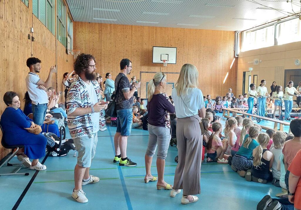 Links sitzen oder stehen Familienangehörige, dann eine Reihe von Lehrer*innen und Erzieher*innen, auf dem Boden sitzen die Schüler*innen und schauen dem Stück zu. Am rechten Rand sehen wir ein paar Kinder mit gelben T-Shirts, die zur „unterdrückten“ Gruppe gehören.