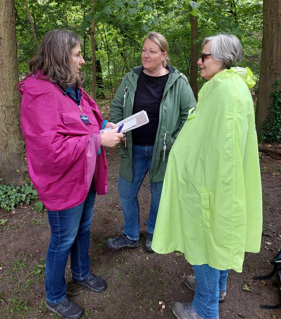 Die drei Frauen des Tridems stehen unter grünen Bäumen und unterhalten sich, die Koordinatorin hält Unterlagen in der Hand und hat einen pinken Regenponcho an, Susanne auf der rechten Seite einen hellgrünen.