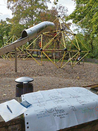 Das Foto zeigt im Vordergrund eine Mauer, auf der ein Plakatentwurf, Schreibzeug und ein Kühlpad liegen. Hinter der Mauer steht ein Mülleimer und im Hintergrund ist der Spielplatz mit einem Klettergerüst, einer Rutsche und herbstlich gefärbte Bäume.