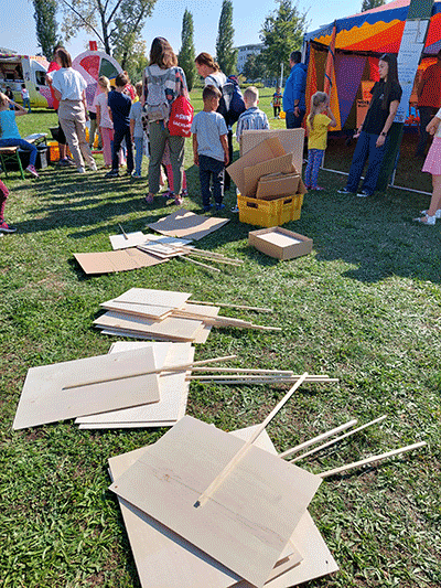 Im Vordergrund liegen blanko Holzplatten mit Stiel, die als Plakate gestaltet werden können. Im Hintergrund sieht man ein buntes Zelt und eine Schlange von Kindern, die bei einem Glücksrad für das Klimaspiel anstehen.