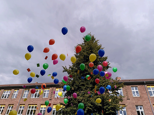 Viele bunte Ballons steigen in den wolkigen Himmel, im Hintergrund sieht man das Schulgebäude und die große Tanne vom Schulhof.