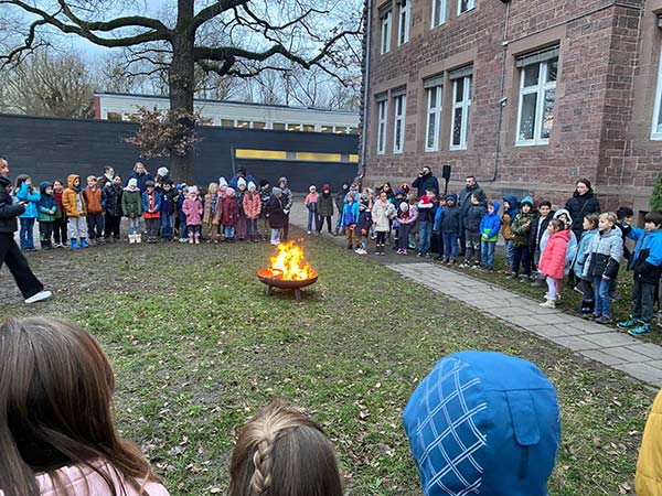 Ein großer Kreis von warm angezogenen Kindern und Lehrer*innen steht um eine Feuerschale herum auf dem Rasenteil des Schulhofs zum gemeinsamen Singen.