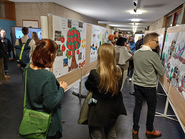 In einem Gang der Hochschule stehen Pinwände mit Postern oder große Bildschirme mit Präsentationen, die von den Teilnehmer*innen der Tagung angeschaut und besprochen werden.)