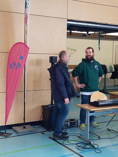 In der Turnhalle stehen auf der linken Bildseite mobile Fahnen des stja und der Schillerschule. Vor einem Tisch mit einem Keyboard unterhält sich der stja Geschäftsführer Daniel Melchien (links) mit dem Schulleiter Frank Hutt.