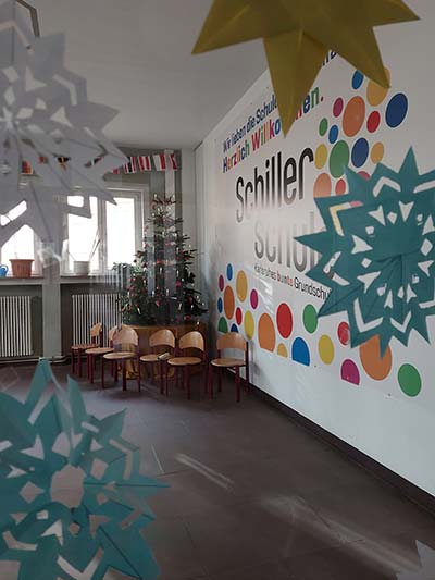 Durch eine Glastür, die mit Papiersternen dekoriert ist, sieht man den Eingangsbereich der Schule, auf der linken Wand ist groß das Schullogo angebracht, in der Ecke steht ein Weihnachtsbaum.